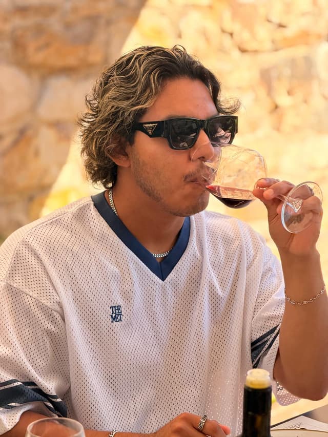 Guest tasting a glass of red wine against the sunlit stone wall of the winery
